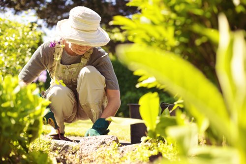 A woman wearing a wide-brimmed hat, glasses, and gardening gloves is kneeling on a well-maintained lawn in a backyard garden, engaged in planting or tending to flower beds. The garden features a lush, green lawn with dense, leafy plants and shrubbery along the borders. In the background, there are mature trees with a canopy of leaves providing shade, and sunlight filters through, creating a bright, warm outdoor environment typical of a well-kept garden in Beckenham. The gardener is focused on her tasks, with gardening tools nearby, and the scene highlights the natural textures of soil, grass, and foliage, supporting gardening services such as lawn care, planting, and outdoor maintenance in the area around postcode BR3 or BR4. The overall setting conveys a peaceful, tidy external space suitable for horticultural activities and garden enhancement.