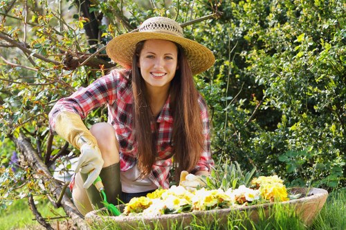 A young woman with long brown hair, wearing a wide straw hat, a red and white checked shirt, and gardening gloves, is kneeling in a lush garden surrounded by greenery and flowering plants. She is smiling and holding a small gardening tool, while tending to a large, overflowing planter filled with vibrant yellow and purple flowers. The garden features a well-maintained lawn with dense shrubbery and trees in the background, indicating a typical suburban outdoor space in the Beckenham area. The scene appears to be a sunny day with natural daylight illuminating the garden, emphasizing its healthy plants and neat layout, reflecting professional gardening and horticultural practices typical of local gardening services such as Hedge Trimming Beckenham.