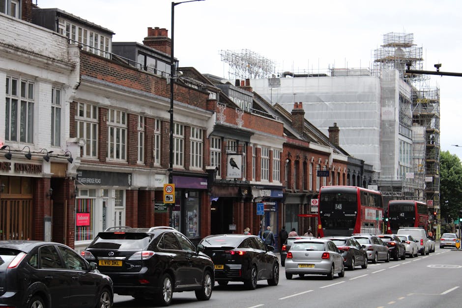 A busy London street scene featuring traditional brick-fronted shops with white-framed windows and signage, parked cars lining the curb, and two red double-decker buses in motion. The buildings are a 