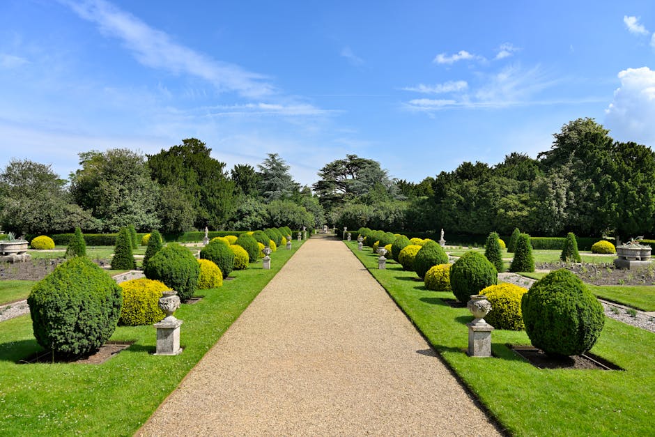 A person wearing a blue raincoat with a hood, black trousers, and safety shoes is engaged in garden maintenance, trimming a hedge with pruning shears in a well-kept garden. The garden features a neatl