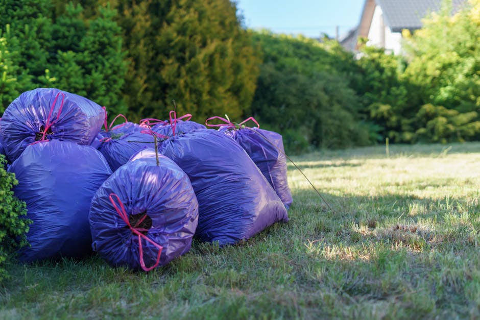 A row of tightly packed purple garden waste bags filled with trimmed hedge clippings and yard debris, tied with pink strings, are placed on a well-maintained grassy lawn. The garden is bordered by den