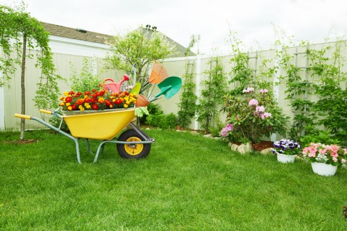 A well-maintained backyard garden featuring a lush, green lawn with evenly cut grass, bordered by a white wooden fence adorned with climbing plants and vines. In the foreground, a yellow garden wheelbarrow is filled with colourful flower pots, gardening tools, and decorative items, indicating recent garden care or planting. To the right, there are flower beds with blooming pink, purple, and red flowers, arranged in neat borders along the fence, with some in white pots placed on the grass. A small shrub and a young tree stand behind the flower beds, adding height and variety to the garden layout. The environment appears bright and clear, suggesting a sunny day with natural daylight illuminating the garden, which is typical of outdoor spaces maintained by professional gardening services such as Hedge Trimming Beckenham, especially around the postcode area of Beckenham in South London. Overall, the scene depicts a tidy, vibrant outdoor space ready for relaxing or gardening activities, reflecting professional gardening and lawn care standards.