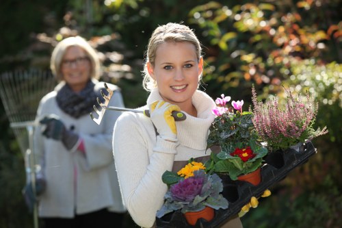 A young woman in a cream sweater and gardening gloves is smiling and holding a black tray with pink, purple, and yellow flowering plants, highlighting her gardening activity outdoors in a garden setting. In the background, an older woman with glasses, wearing a white jacket and a scarf, is slightly blurred, also engaged in gardening. The garden features a lawn with dense, well-maintained grass in the foreground, surrounding shrubs and vibrant foliage in various shades of green, red, and orange, indicative of seasonal change. Sunlight filters through the trees, creating a warm natural glow that illuminates the scene. The background includes a mix of bushes and taller trees, contributing to a lush outdoor environment typical of a landscaped garden or backyard in Beckenham or South London. The setting emphasizes outdoor plant care and garden maintenance, aligning with professional gardening services offered by Hedge Trimming Beckenham. The scene captures a moment of gardening activity, with natural textures of grass, leaves, and flowers clearly visible, illustrating a vibrant and healthy garden environment.
